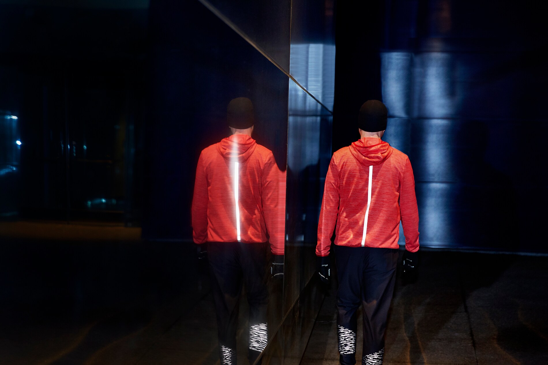 Illuminated Black Stretch Reflective stripe down back of running jacket on man walking next to wall of mirrors at night.