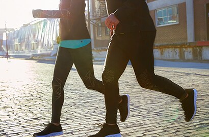 Man and woman running on cobblestone street at sunset wearing running tights with 3M Scotchlite Black Stretch Reflective.