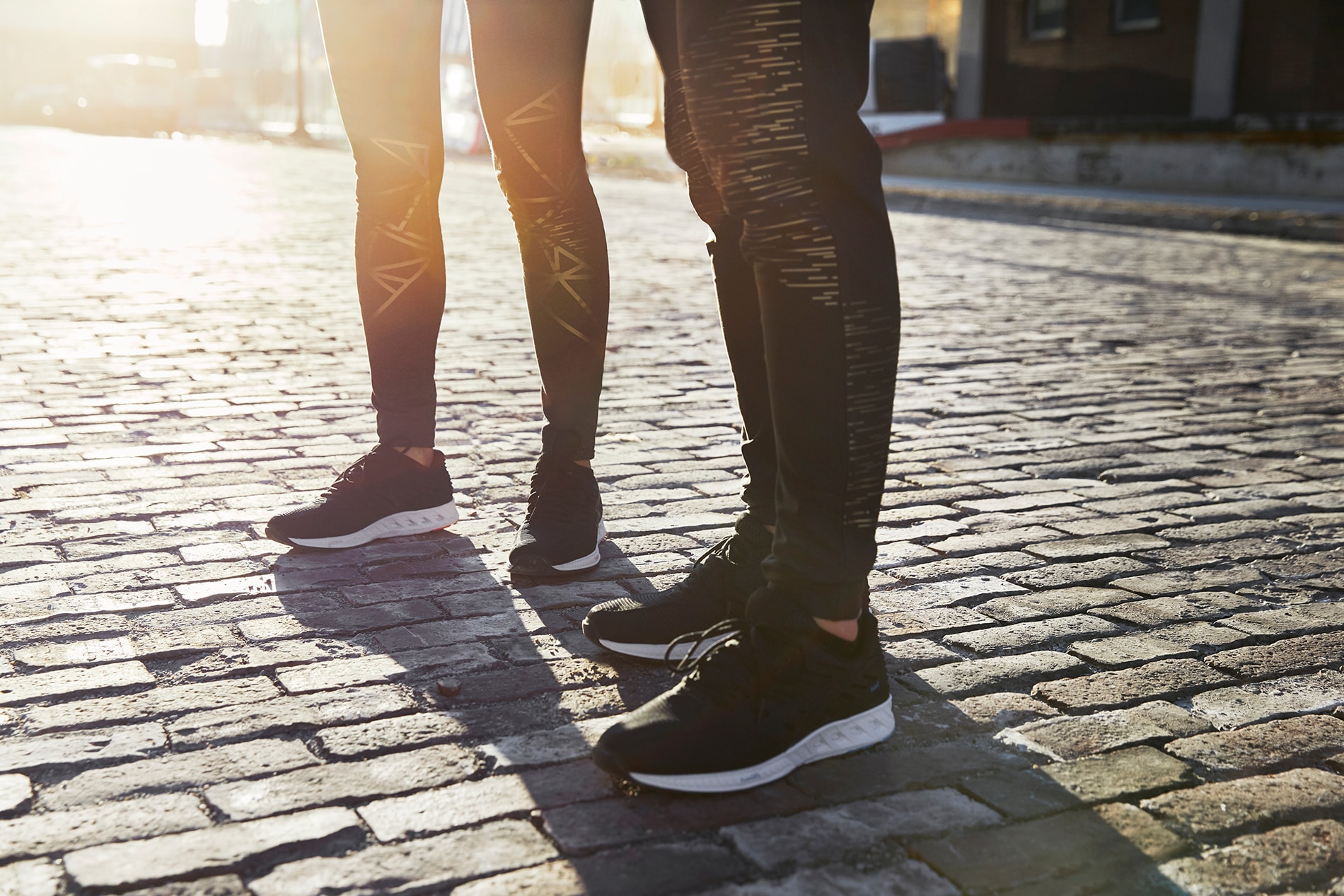 Man and woman standing on cobblestone street at sunset wearing running tights with 3M Scotchlite Black Stretch Reflective.

