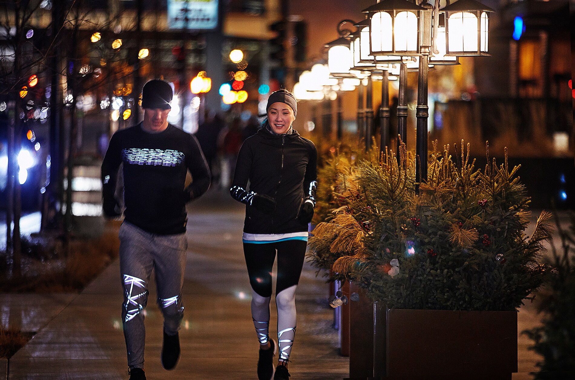 Man and woman running under city lights wearing running gear illuminated by 3M Scotchlite Black Stretch Reflective.
