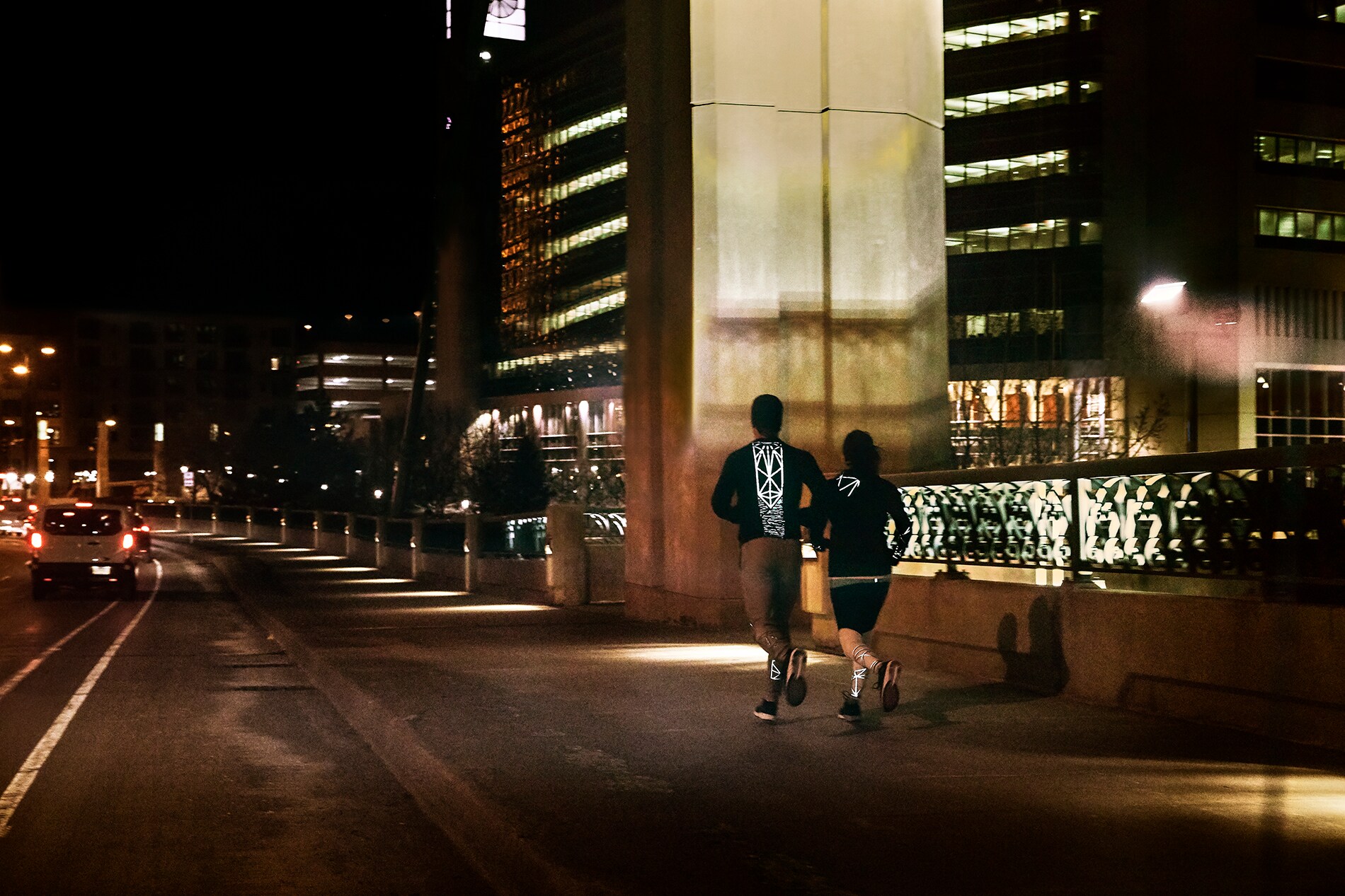 Man and woman running across city bridge wearing running gear illuminated by 3M Scotchlite Black Stretch Reflective.
