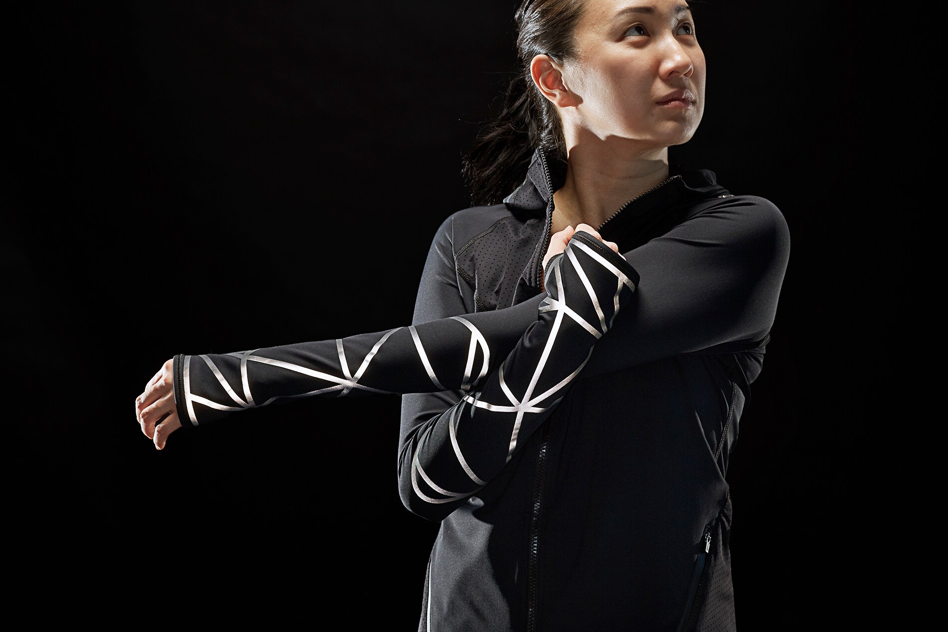 Woman stretching arms wearing jacket, pants illuminated by 3M Scotchlite Black Stretch Reflective.