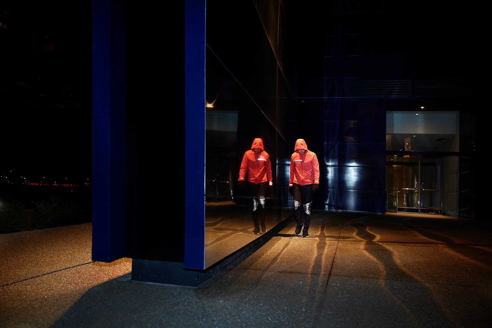 Illuminated Black Stretch Reflective knee panels on running tights on man walking next to wall of mirrors at night.
