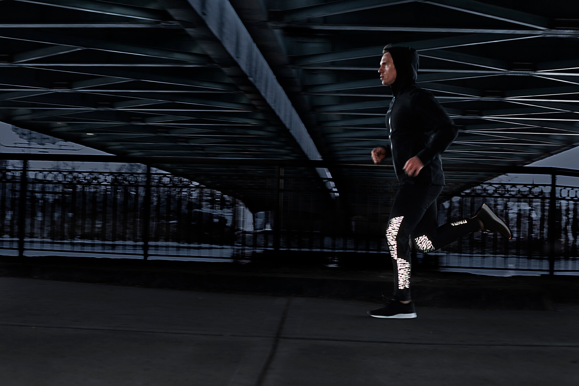 Man running under city bridge at dusk wearing running tights illuminated by 3M Scotchlite Black Stretch Reflective.
