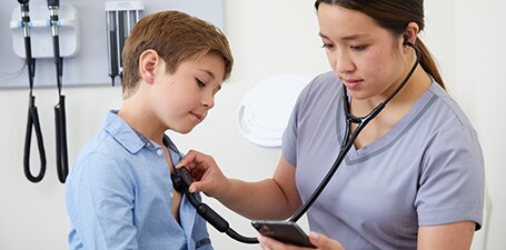 Médico ausculta o peito de um rapaz enquanto olha para a aplicação no telemóvel
